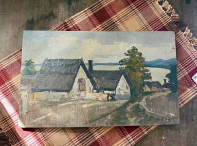 An oil painting of a rural landscape with thatched-roof cottages, a tree, and a winding road, displayed on a red and beige plaid blanket over a wooden surface.