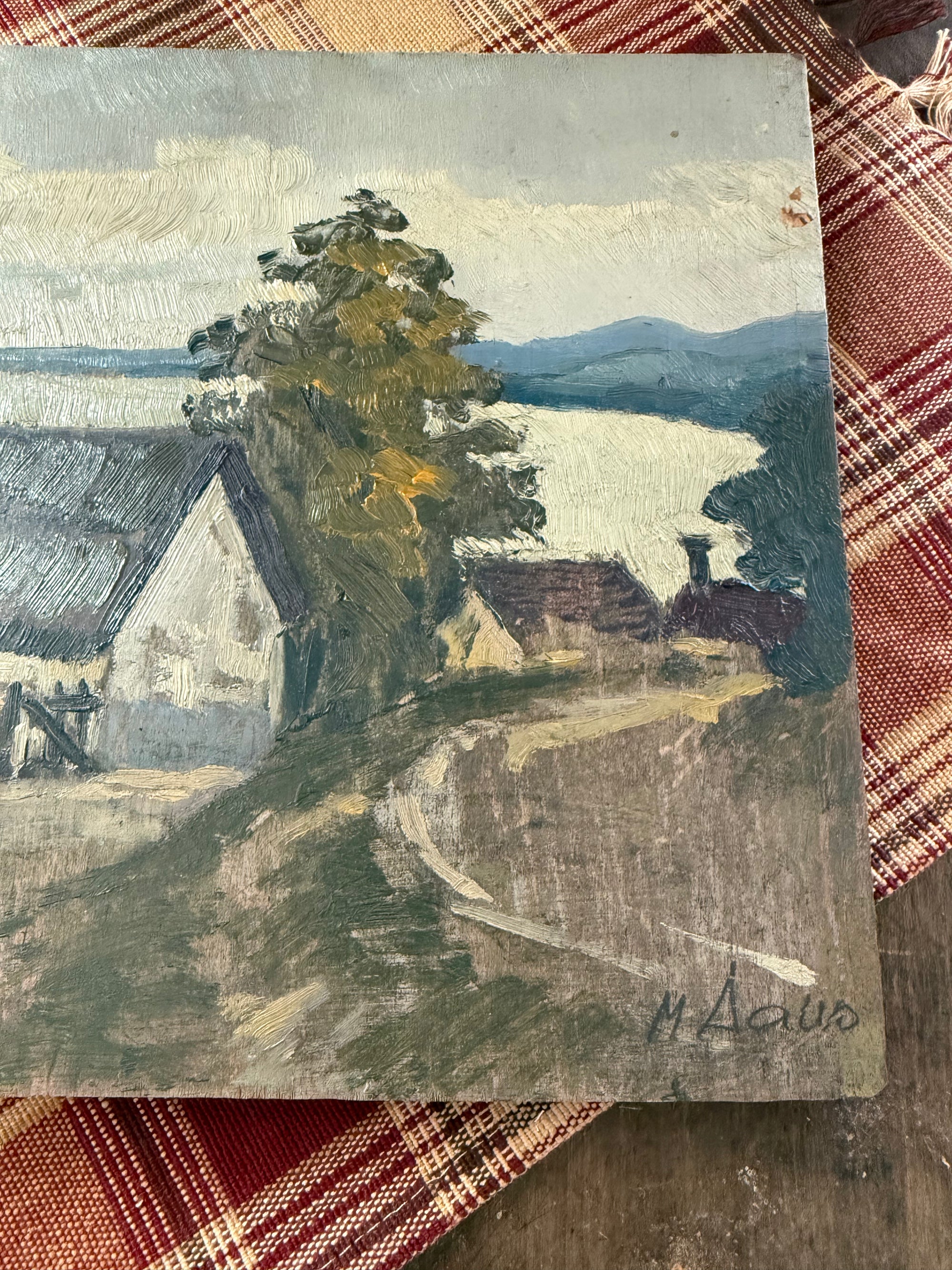 An oil painting of a coastal landscape with houses, a large tree, a winding path, and distant mountains over water, signed 'M. Dalo' in the bottom right corner, displayed on a red and white plaid fabric surface.