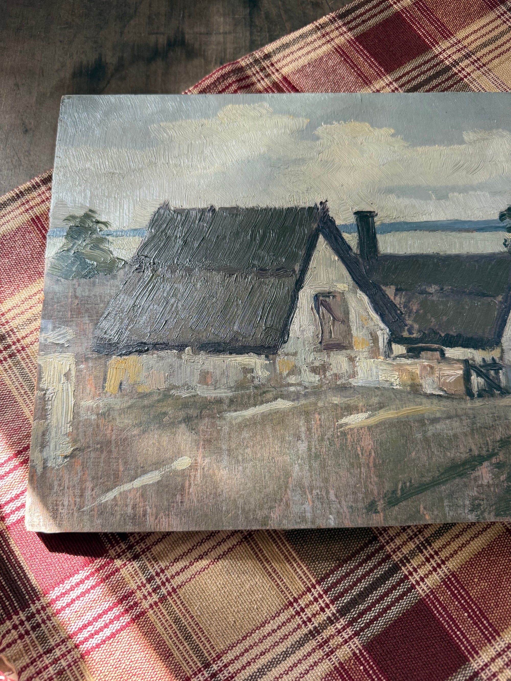 A square canvas painting depicting a rustic cottage with a thatched roof, resting on a red and beige plaid fabric surface.