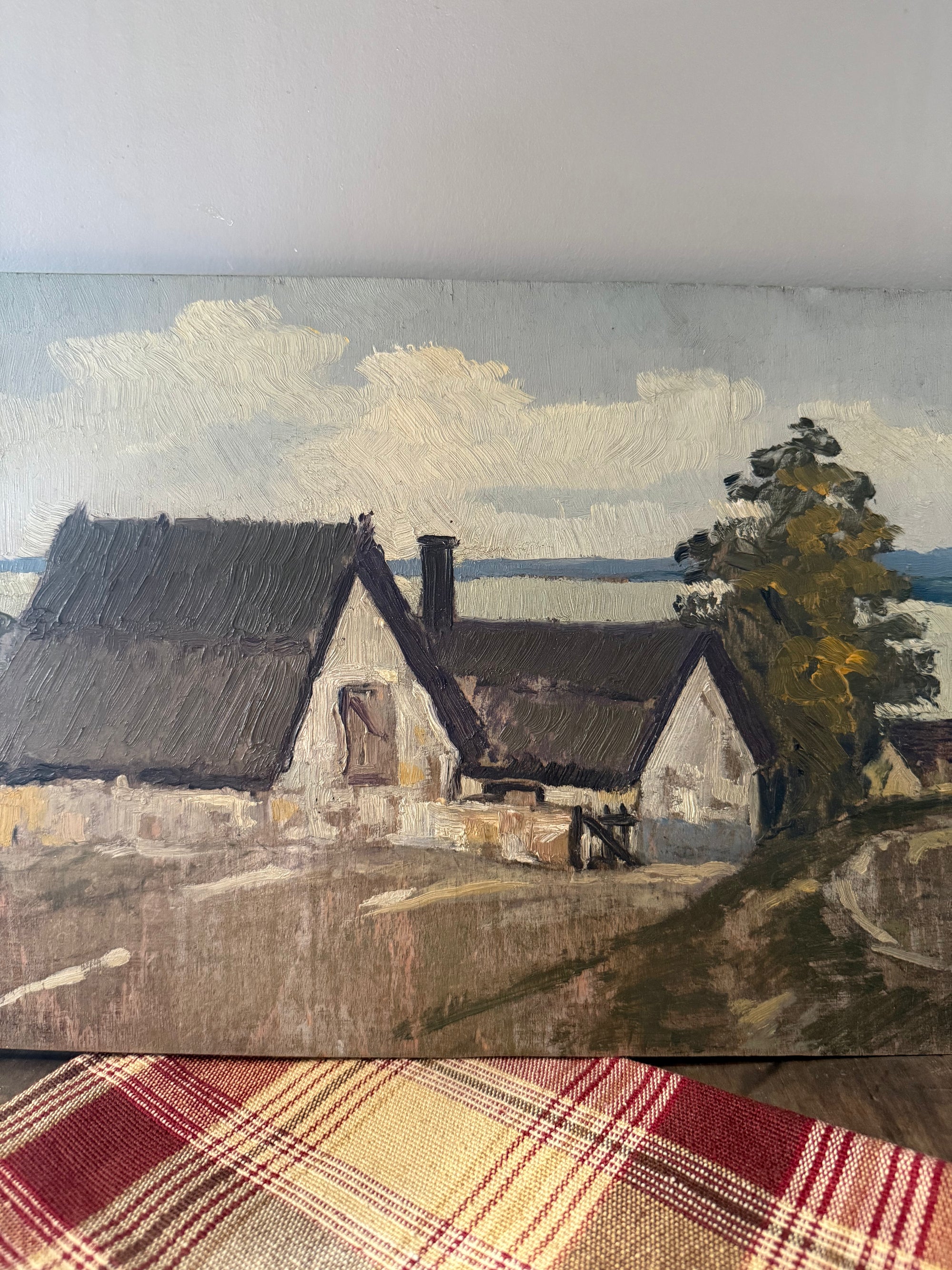 An oil painting of a rural landscape with two cottages, a tree, and a cloudy sky, displayed on a plaid blanket over a wooden surface.