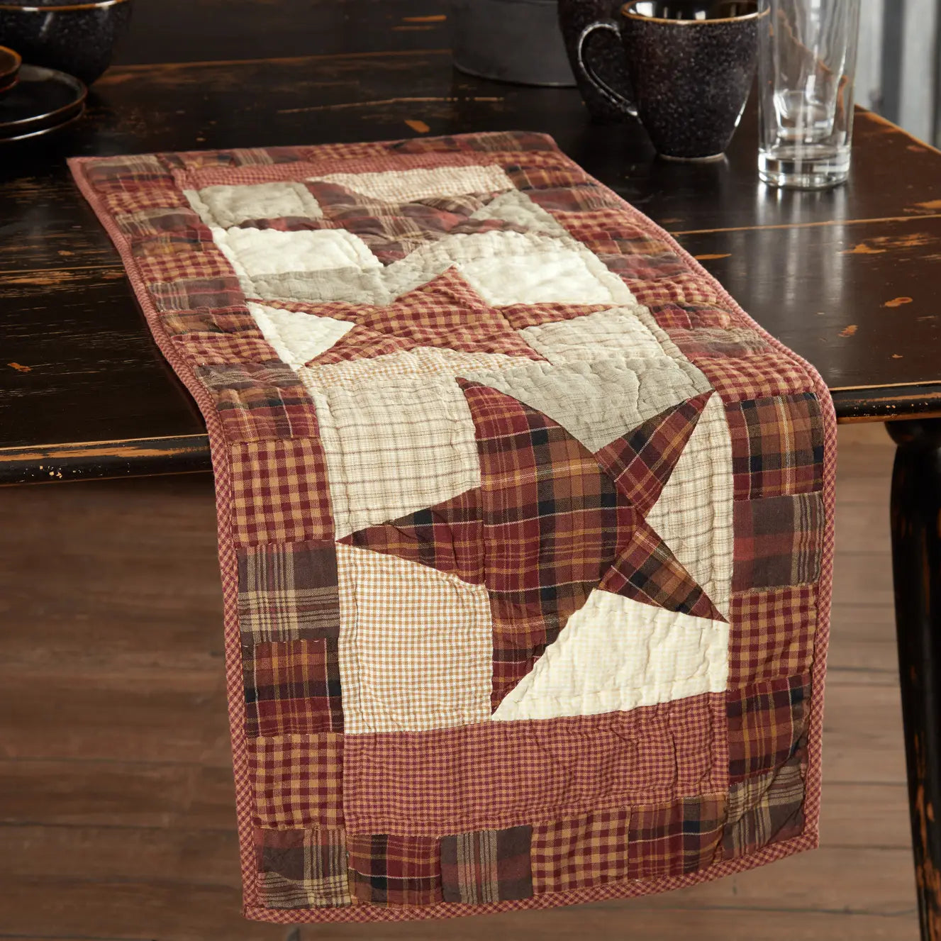 A rustic quilted table runner with a patchwork design featuring red, brown, and cream stars and plaid borders, displayed on a dark wooden dining table.