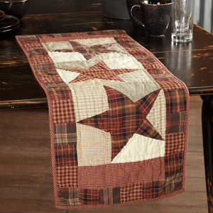 A rustic quilted table runner with a patchwork design featuring red, brown, and cream stars and plaid borders, displayed on a dark wooden dining table.