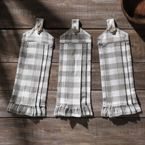 Three gray and white buffalo plaid kitchen towels with ruffled hems and snap tabs, displayed on a rustic dark wood plank background.