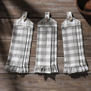 Three gray and white buffalo plaid kitchen towels with ruffled hems and snap tabs, displayed on a rustic dark wood plank background.