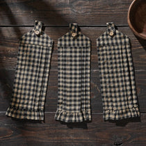 Three matching plaid kitchen towels with ruffled hems hang on a dark wooden surface. The towels feature a black and beige gingham pattern, each with a small wooden button at the top and a ruffled hem at the bottom.