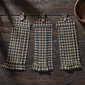 Three matching plaid kitchen towels with ruffled hems hang on a dark wooden surface. The towels feature a black and beige gingham pattern, each with a small wooden button at the top and a ruffled hem at the bottom.