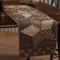 A rustic quilted table runner with a patchwork design in brown, beige, and black gingham fabrics, draped over a dark wooden dining table.