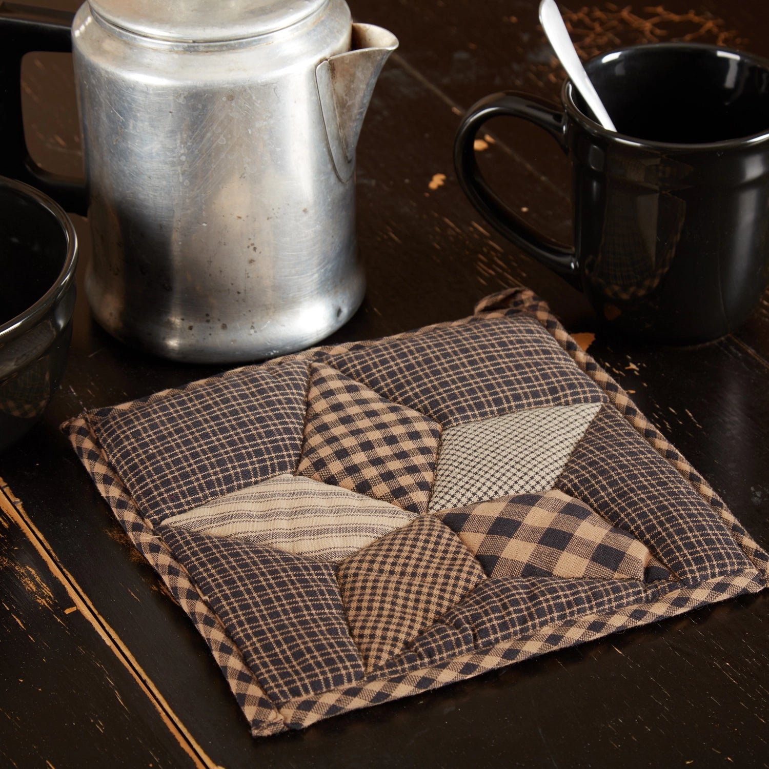 A square quilted fabric trivet or coaster with a patchwork star design in brown, beige, and black gingham patterns, styled on a dark wooden table next to a metal coffee pot, a black mug, and a bowl.