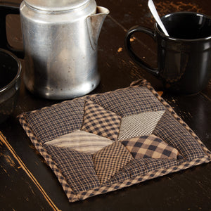 A square quilted fabric trivet or coaster with a patchwork star design in brown, beige, and black gingham patterns, styled on a dark wooden table next to a metal coffee pot, a black mug, and a bowl.
