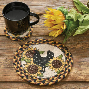 Two round braided coasters on a rustic wooden table, one featuring a black rooster and the other a sunflower design, with a black coffee mug and yellow sunflowers placed on top.
