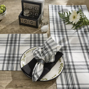 A styled tabletop setting featuring black and white plaid and striped table linens, including a place setting with a white plate, black napkin, and matching napkin ring, alongside a salt and pepper holder and a white flower on a wooden table.