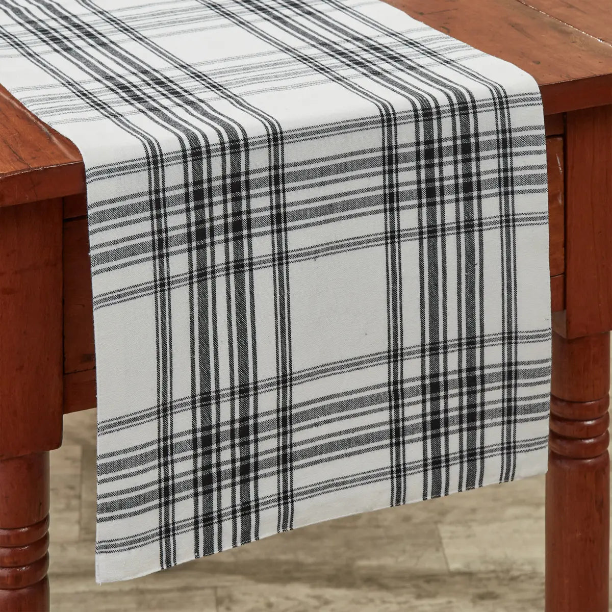 A white table runner with a black plaid pattern draped over a rustic reddish-brown wooden table.