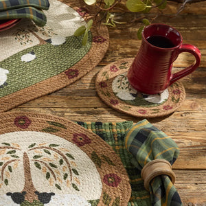 A styled tabletop setting featuring woven placemats and coasters with a tree and floral pattern, a red ceramic mug, and a green plaid napkin on a rustic wooden table.