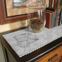 A light gray embroidered lace runner with a floral and leaf pattern laid across a dark wooden dresser, with a clear glass jar and books as props.
