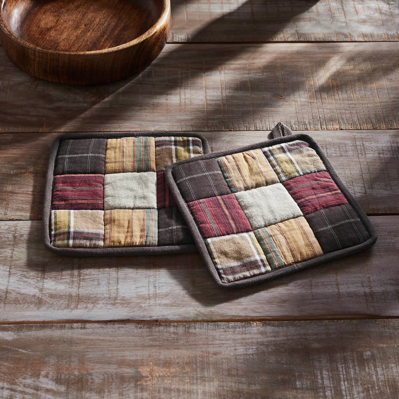 Two rectangular patchwork potholders with a rustic plaid and stripe pattern in earthy tones, displayed on a weathered wooden tabletop next to a wooden bowl.