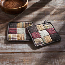 Two rectangular patchwork potholders with a rustic plaid and stripe pattern in earthy tones, displayed on a weathered wooden tabletop next to a wooden bowl.