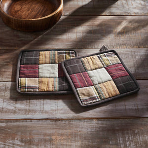 Two rectangular patchwork potholders with a rustic plaid and stripe pattern in earthy tones, displayed on a weathered wooden tabletop next to a wooden bowl.